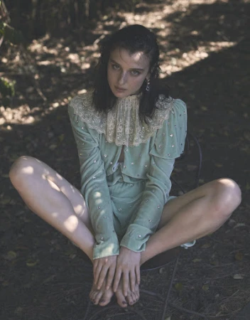 A light-skinned woman with dark hair is sitting barefoot outdoors in a chair wearing a light green embellished outfit.