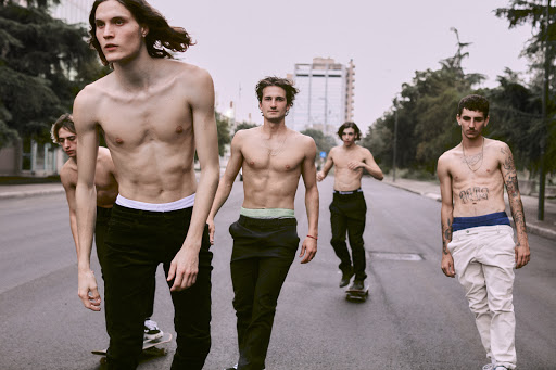 Group of five shirtless young men walking with skateboards on an asphalt road in front of a tall building and some trees.