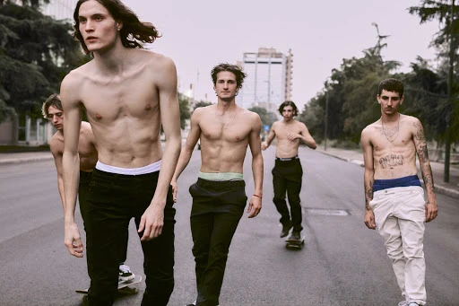 Group of five shirtless young men walking with skateboards on an asphalt road in front of a tall building and some trees.