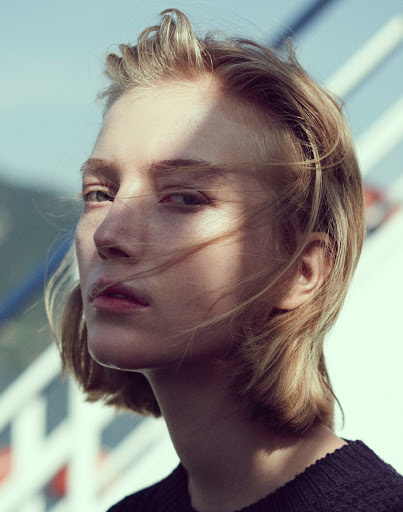 Close-up portrait of a blonde adult woman with short hair and fair skin, outside in natural light, wearing a dark sweater, against a light blue sky.