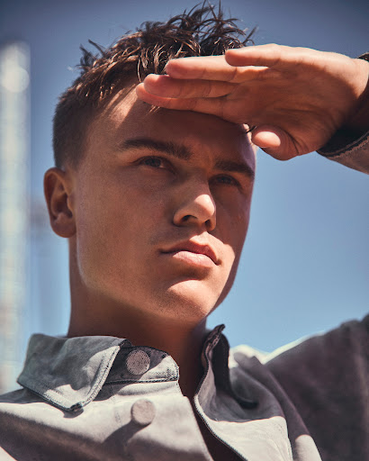 Portrait of a young adult male shielding his eyes with his hand in an outdoor setting with buildings and a clear blue sky.