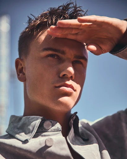 Portrait of a young adult male shielding his eyes with his hand in an outdoor setting with buildings and a clear blue sky.