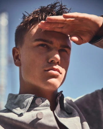 Portrait of a young adult male shielding his eyes with his hand in an outdoor setting with buildings and a clear blue sky.