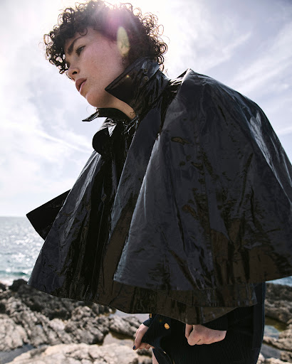 Woman in glossy black cape stands on rocks by the sea under bright sunlight. Fashion shot with natural light.