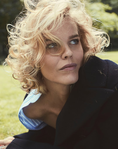 Close-up portrait of a woman with blonde curly hair and blue eyes wearing a black jacket outdoors.