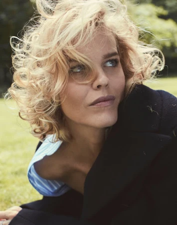 Close-up portrait of a woman with blonde curly hair and blue eyes wearing a black jacket outdoors.