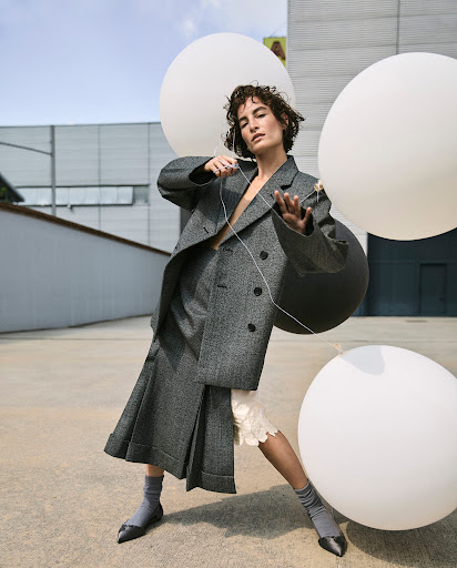 Fashion editorial shot of a woman in a gray suit, posing with white balloons, against an urban background. Modern fashion and minimalist style.