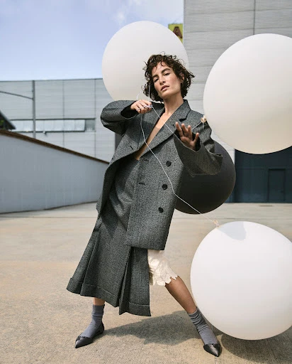Fashion editorial shot of a woman in a gray suit, posing with white balloons, against an urban background. Modern fashion and minimalist style.