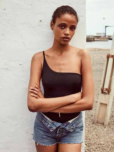 Woman in a black bodysuit and denim shorts poses against a white wall with a neutral expression, outdoors, on a sunny day near a coastal area.