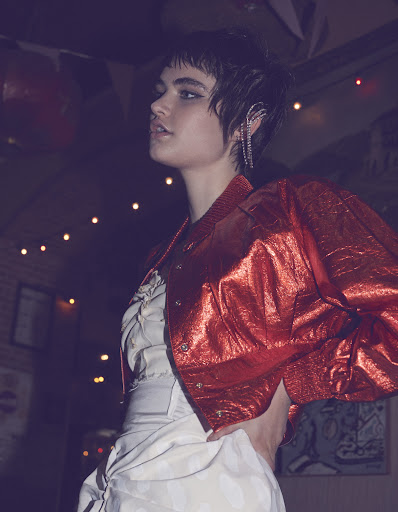 A young adult woman with short hair poses in a metallic red jacket and a white dress with a dark, blurred background.