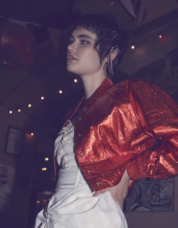 A young adult woman with short hair poses in a metallic red jacket and a white dress with a dark, blurred background.