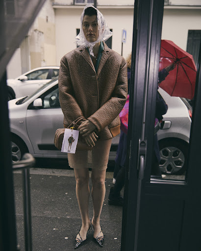 Fashionable adult in a tan blazer, clear head covering, skirt, and handbag, standing in front of a storefront on a city street.