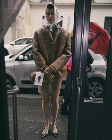 Fashionable adult in a tan blazer, clear head covering, skirt, and handbag, standing in front of a storefront on a city street.
