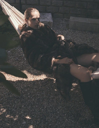 Adult woman in a fur coat reclines in a hammock outdoors on a stone patio, with a brick wall in the background, and green foliage.