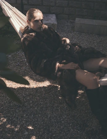 Adult woman in a fur coat reclines in a hammock outdoors on a stone patio, with a brick wall in the background, and green foliage.
