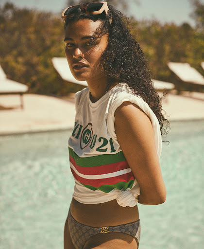Outdoor portrait of a young adult woman with sunglasses on her head, wearing a printed tee and bikini bottoms, with a pool behind her.