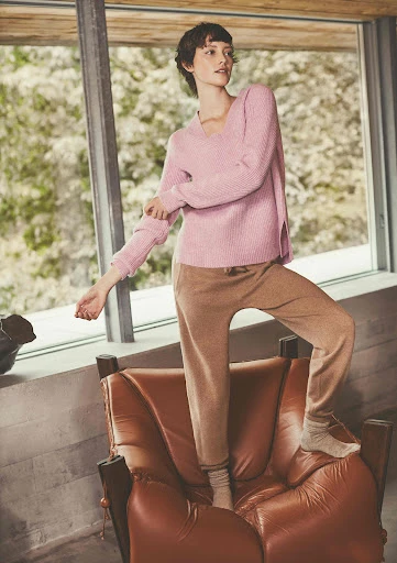 Adult female in a pink sweater and tan sweatpants standing on a brown leather chair indoors near a window with outdoor foliage. 