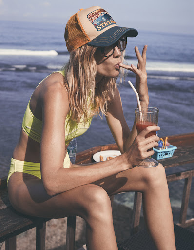 Woman in yellow bikini and Stetson cap sits at a table by the sea, enjoying a cocktail with a straw and holding up her finger.