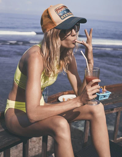Woman in yellow bikini and Stetson cap sits at a table by the sea, enjoying a cocktail with a straw and holding up her finger.