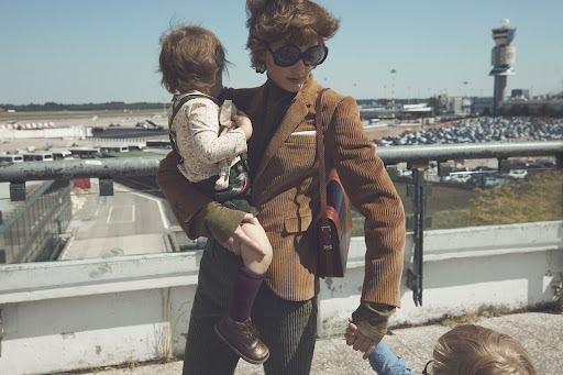 Stylish woman in a corduroy blazer holds a young child, standing near an airport with an observation tower, evoking a sense of travel.
