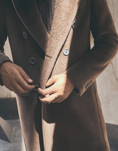Close-up of an adult male buttoning a brown double-breasted wool overcoat, showcasing fashion, style, and texture against a neutral background.