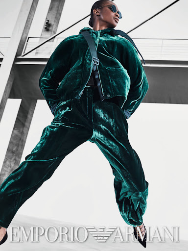 Fashion editorial shot of a Black woman in a green velvet suit with a cross-body bag, posing against a modern architectural background.