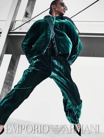 Fashion editorial shot of a Black woman in a green velvet suit with a cross-body bag, posing against a modern architectural background.