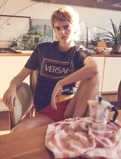 Stylish model sits in chair wearing a Versace T-shirt, captured indoors with natural light, alongside a Moka pot and tablecloth.