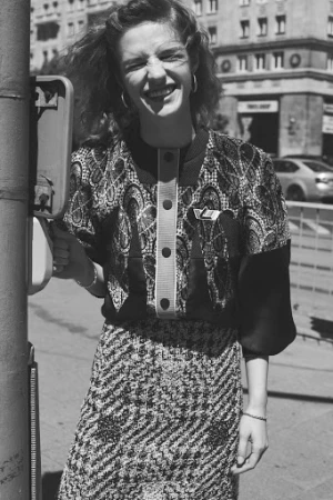 Smiling woman wearing a patterned patterned button-down top and tweed skirt outdoors in a city street.