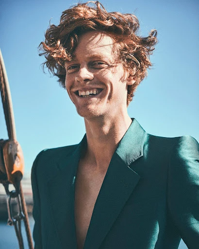 Smiling redhead man in a dark green blazer standing on a sailboat, with blue sky and harbor in the background.