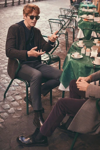 Two men seated at an outdoor cafe in Rome, Italy. One man gesturing, wearing a blazer, the other is partially visible.
