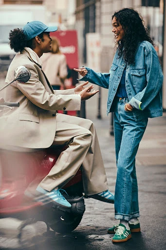 Two stylish adults in denim and tan blazers stand next to a red moped on a city street, smiling and gesturing in daylight.