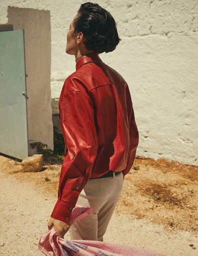 Adult in red leather shirt looks up next to white wall in outdoor shot; holding pink and blue fabric in hand in daylight.