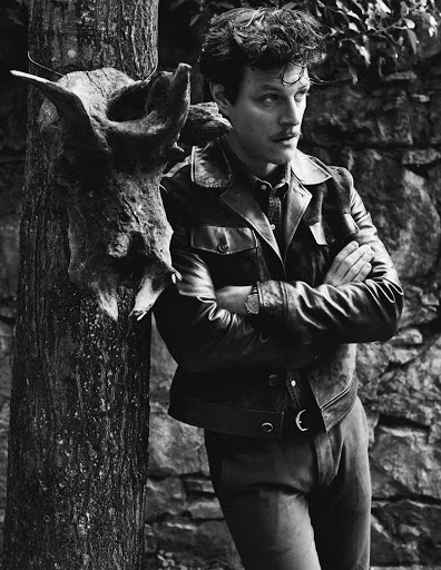 Monochrome portrait of a man with a mustache leaning against a tree with a deer skull, wearing a leather jacket and collared shirt.