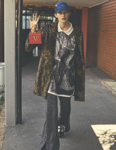 Fashionable woman in leopard coat, graphic tee, blue cap, and wide leg pants walking in an urban tunnel with a small red purse.