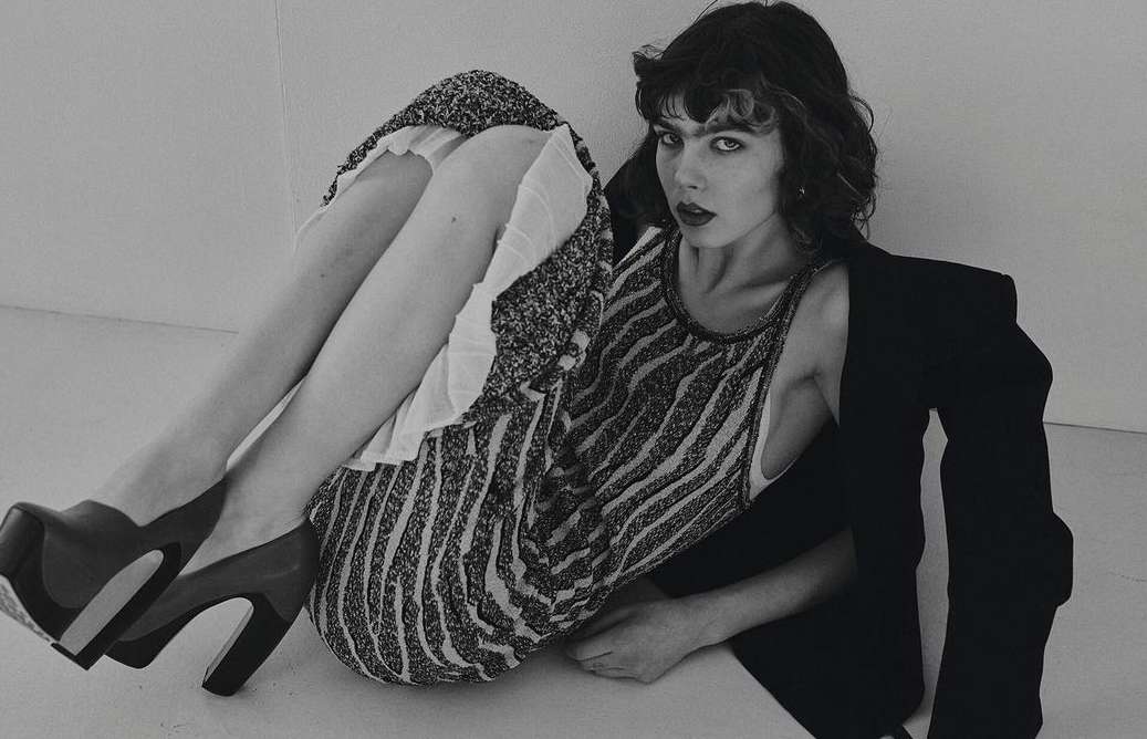 Model with dark curled hair wearing a patterned dress and platform heels posing against a plain background
