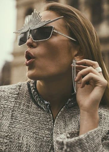 Blonde woman poses in embellished sunglasses and grey tweed jacket with Roma cityscape backdrop; bold fashion statement.
