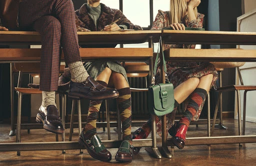 Three models seated at wooden desks wearing patterned socks and leather shoes, photographed by Stefano Galuzzi.