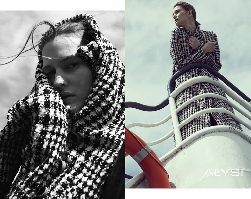 Monochrome outdoor fashion shot: woman with patterned coat, windblown hair, and life preserver visible against a partly cloudy sky.