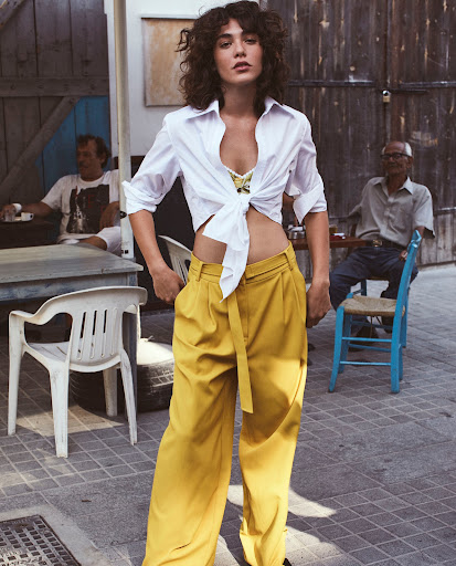 Woman in a white shirt and yellow pants standing in front of an outdoor building with two men sitting in the background.