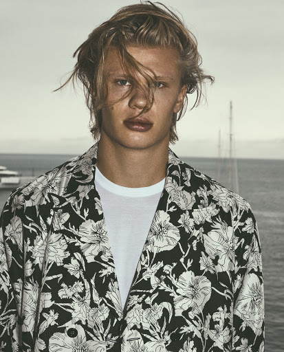 Blond male model in a floral button-down shirt stands outdoors with yachts in the background, looking directly at the camera.