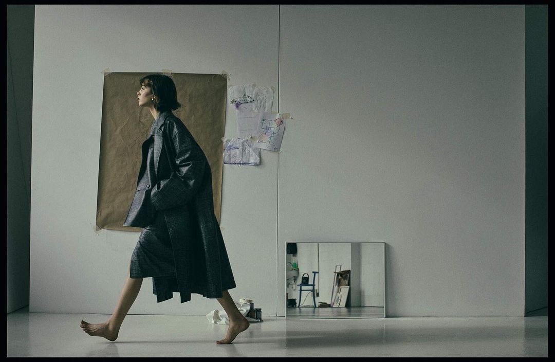 A barefoot woman in a gray coat walks through a minimalist space near a mirror. A brown paper sheet is hung on the wall behind her.