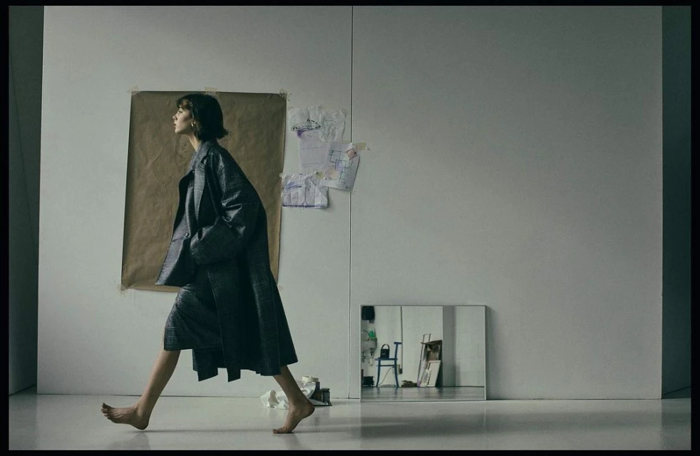 A barefoot woman in a gray coat walks through a minimalist space near a mirror. A brown paper sheet is hung on the wall behind her.