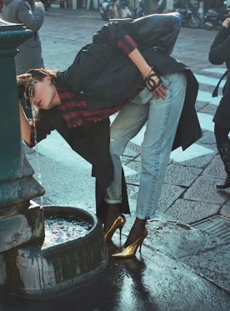 A fashionable young woman drinks from an old public fountain outdoors in Europe, wearing a black coat, jeans, and gold high heels.
