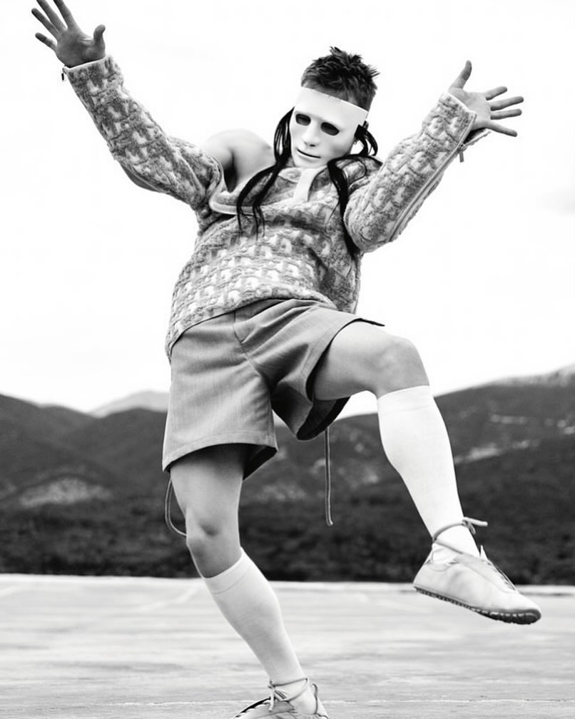 Person in white mask and patterned sweater strikes a dynamic pose against a blurred mountain backdrop, monochrome fashion photography.