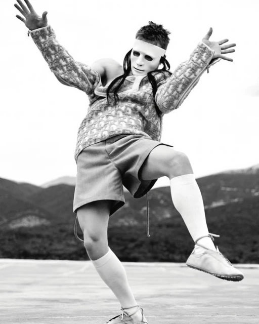 Person in white mask and patterned sweater strikes a dynamic pose against a blurred mountain backdrop, monochrome fashion photography.