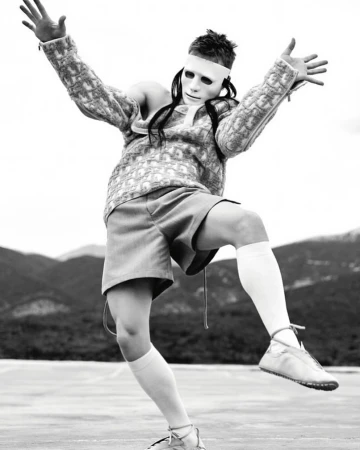 Person in white mask and patterned sweater strikes a dynamic pose against a blurred mountain backdrop, monochrome fashion photography.