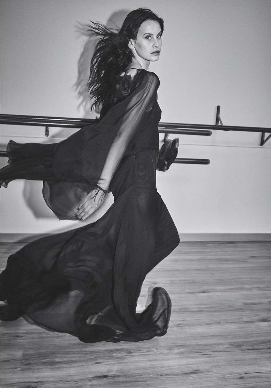 Black-and-white portrait of a young adult woman in a flowing dark dress, walking in front of a barre in a dance studio.