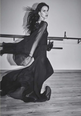 Black-and-white portrait of a young adult woman in a flowing dark dress, walking in front of a barre in a dance studio.