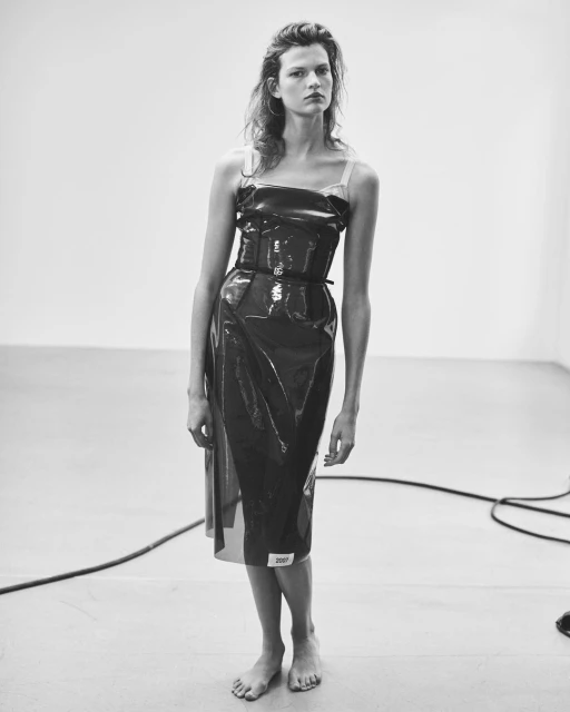 Monochrome portrait of a barefoot female fashion model wearing a dark glossy dress in a photo studio, with a white background.